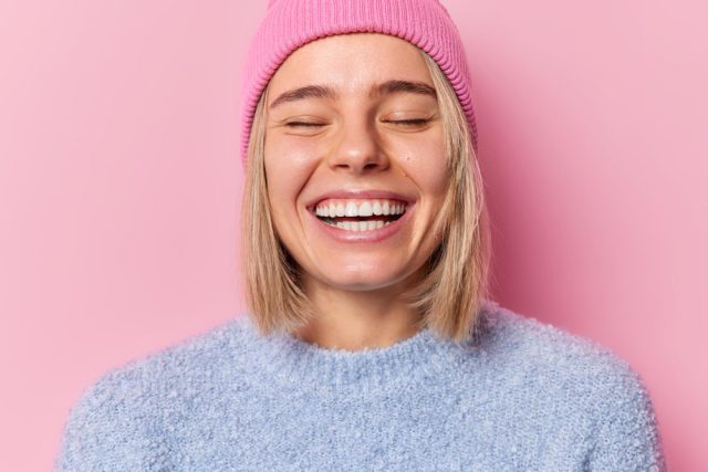 headshot optimistic woman keep eyes closed smile broadly show white teeth expresses sincere feelings feels very glad wears hat casual jumper isolated pink background emotions concept